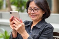 Seorang wanita dengan keterbatasan fisik sedang menggunakan iPhone dengan Hikawa Phone Grip & Stand, tersenyum bahagia.