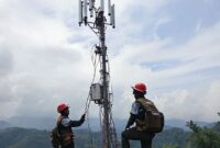 Sebuah tim teknisi Telkomsel bekerja keras di tengah puing-puing pasca bencana, sedang memasang antena Mobile BTS (Combat) di Aceh, dengan latar belakang pemandangan kehancuran dan langit mendung. Mereka mengenakan rompi keselamatan dan helm.