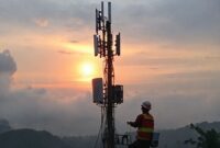 Tim teknisi Telkomsel dengan rompi keselamatan oranye sedang memperbaiki antena BTS yang terendam air banjir di daerah pedesaan Sumatera. Air setinggi pinggang, langit mendung.